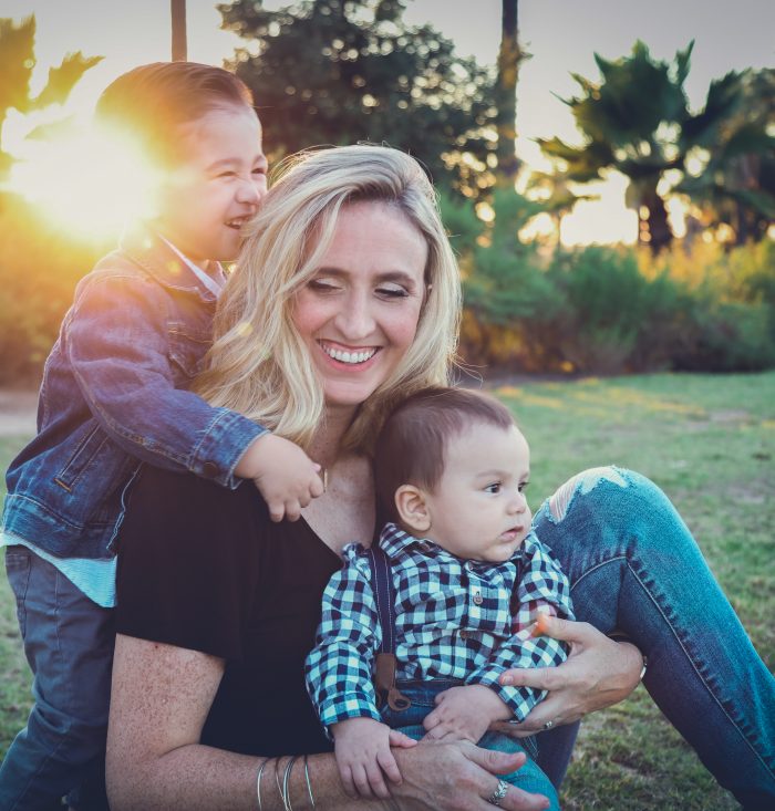 An attractive woman sits on a field during sunset with her two young suns, smiling.