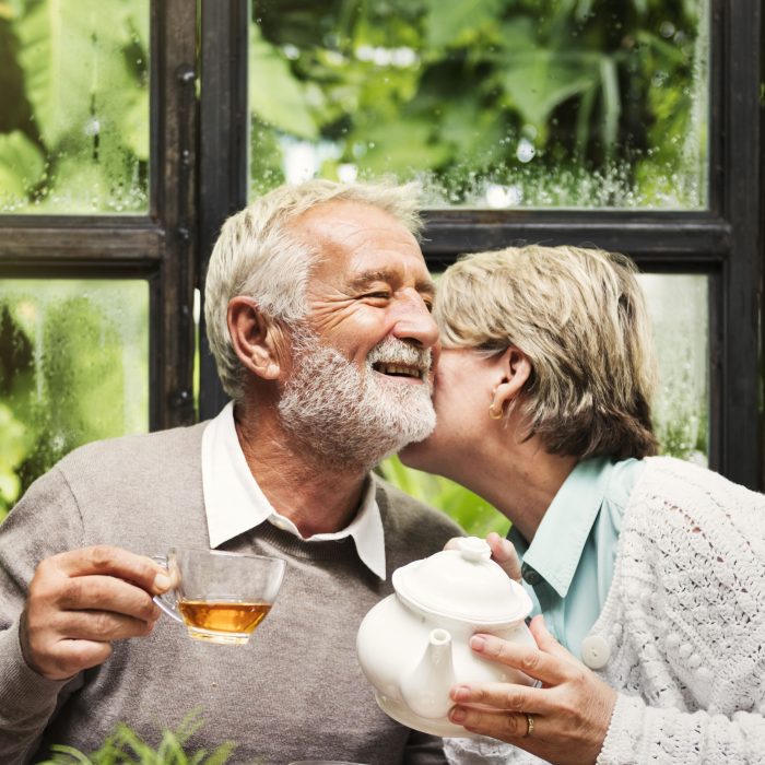 An older couple sit at a table drinking tea as the woman leans over to give the man a kiss on the cheek.