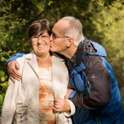 An older couple stand outside and the man kisses the woman on the cheek.