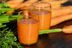 Two large glasses of carrot juice sit on a table in front of carrots.