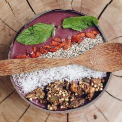 A healthy acai breakfast bowl sits atop a wooden table with a wooden spoon across.